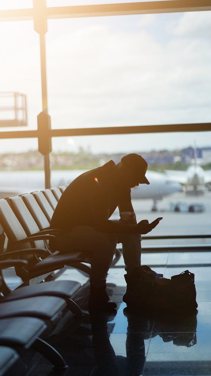 Silhouette of a man using a smartphone in an airport waiting area with airplanes visible outside.